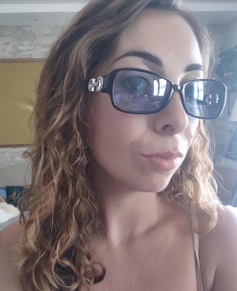 May include: Black rectangular sunglasses with silver embellishments on the temples. The lenses are a gradient purple color. The sunglasses are worn by a person with curly brown hair.