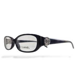 May include: Dark navy blue oval-shaped eyeglasses with clear lenses. The frames feature a silver-toned circular emblem on each temple displaying the interlocking CC logo. The word CHANEL is printed in black on the right lens of the eyewear.