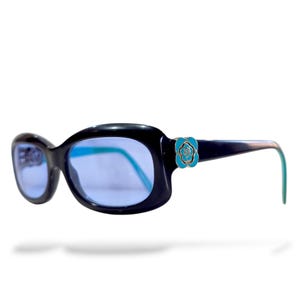 May include: Black oval sunglasses with blue lenses. The frames feature a turquoise flower accent on the temples and turquoise-colored arms. The sunglasses are set against a white background.