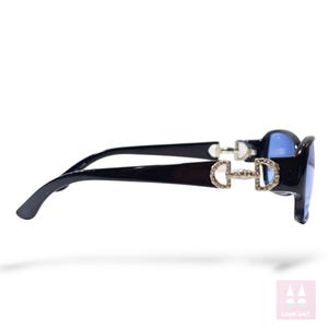 May include: Black sunglasses with blue lenses. The frames feature a decorative silver-colored horse bit design embellished with small, clear rhinestones. The sunglasses have a glossy finish and a curved design.