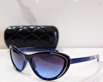 Puede incluir: Gafas de sol azul marino con forma de ojo de gato y lentes degradadas azules. Las monturas están adornadas con ribetes dorados y pequeños adornos. En el fondo, un estuche negro acolchado para gafas de sol. La marca "CHANEL" es visible.
