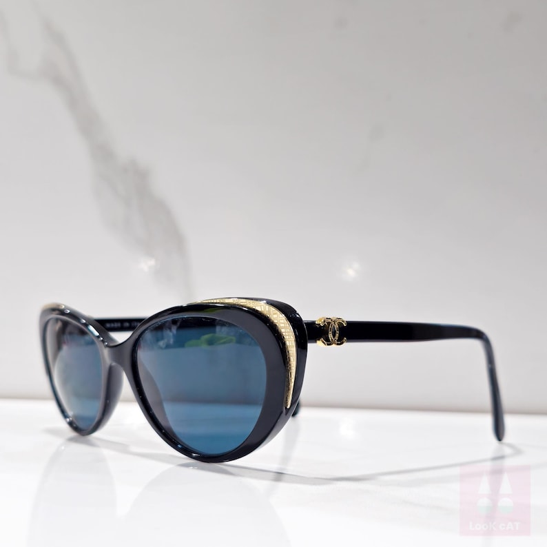 Puede incluir: Gafas de sol negras con forma de ojo de gato y lentes azules. Las monturas presentan detalles dorados y un logotipo dorado en las patillas. Las gafas de sol est&aacute;n sobre un fondo blanco.
