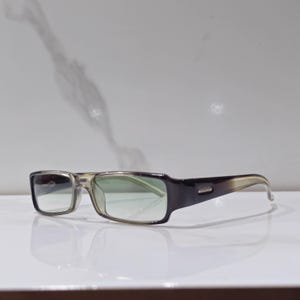 May include: Rectangular eyeglasses with a gradient frame, transitioning from clear to dark brown. The lenses have a green tint. The glasses are set against a white background, reflecting the light.