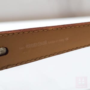 Peut inclure: Gros plan sur une ceinture en cuir marron avec des bords cousus. La ceinture est estampill&eacute;e "roberto cavalli Made in Italy" et la taille "140". La ceinture est de couleur neutre et semble neuve.