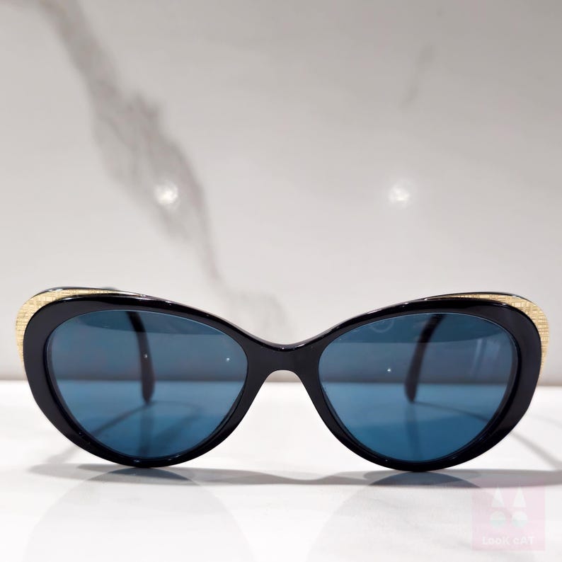 Puede incluir: Gafas de sol negras de ojo de gato con lentes azules. Las monturas son negras con detalles texturizados dorados en las patillas. Las gafas de sol tienen un dise&ntilde;o retro y elegante.