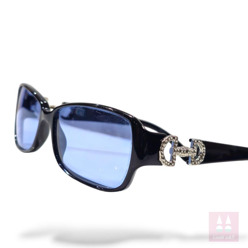 May include: Black sunglasses with blue lenses. The frames have a rectangular shape and are embellished with a silver-colored decorative element on the temples. The lenses are a light blue color.