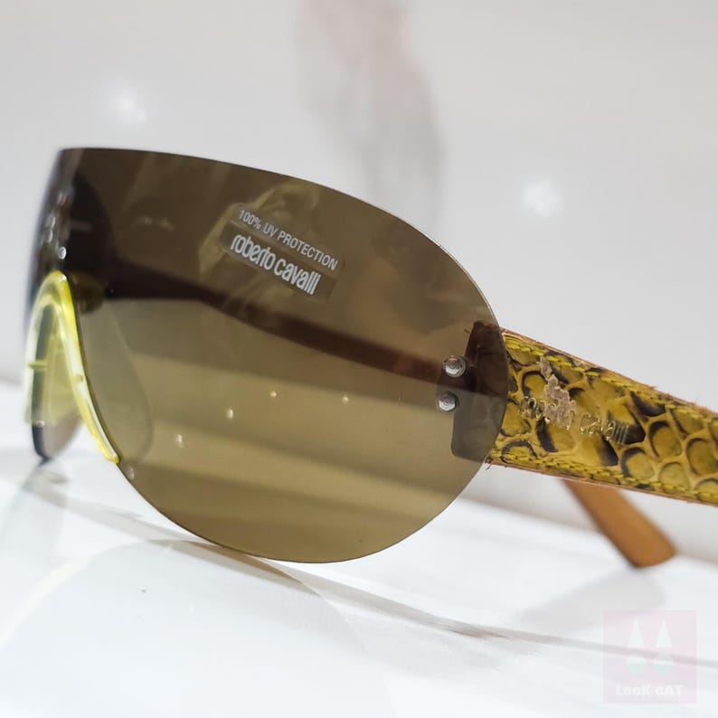 Peut inclure: Lunettes de soleil ovales marron avec des branches &agrave; motif peau de serpent jaune. Les lunettes ont un verre teint&eacute; marron avec le texte "100% UV PROTECTION roberto cavalli".