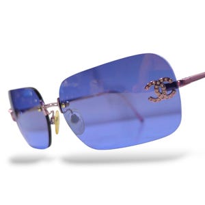 May include: Rimless sunglasses featuring rectangular blue-tinted lenses and thin metallic pink temples. The right lens displays a prominent interlocking CC logo embellished with small clear crystals, complemented by metallic nose pads.
