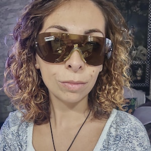 Peut inclure: Lunettes de soleil de style bouclier, teint&eacute;es de marron, avec une monture dor&eacute;e. Les verres refl&egrave;tent l'environnement. La personne porte les lunettes de soleil. L'image montre &eacute;galement un collier avec un pendentif.