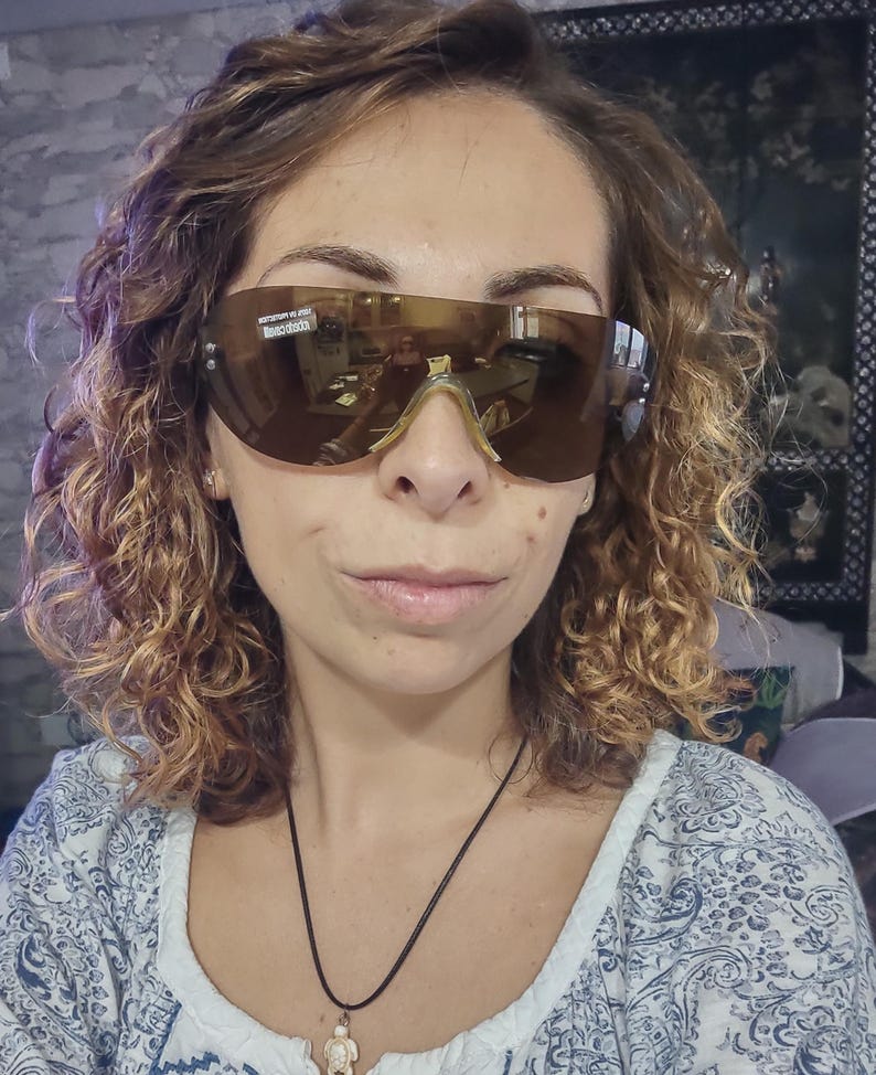 Peut inclure: Lunettes de soleil de style bouclier, teint&eacute;es de marron, avec une monture dor&eacute;e. Les verres refl&egrave;tent l'environnement. La personne porte les lunettes de soleil. L'image montre &eacute;galement un collier avec un pendentif.