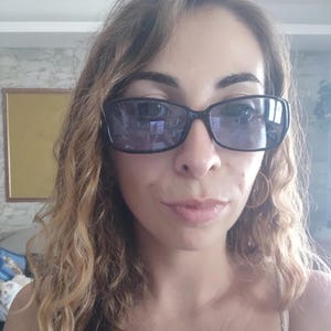 May include: A person wearing black rectangular sunglasses with dark lenses. The person has long, wavy, light brown hair and is wearing gold hoop earrings. The background is a light-colored wall with a framed picture.
