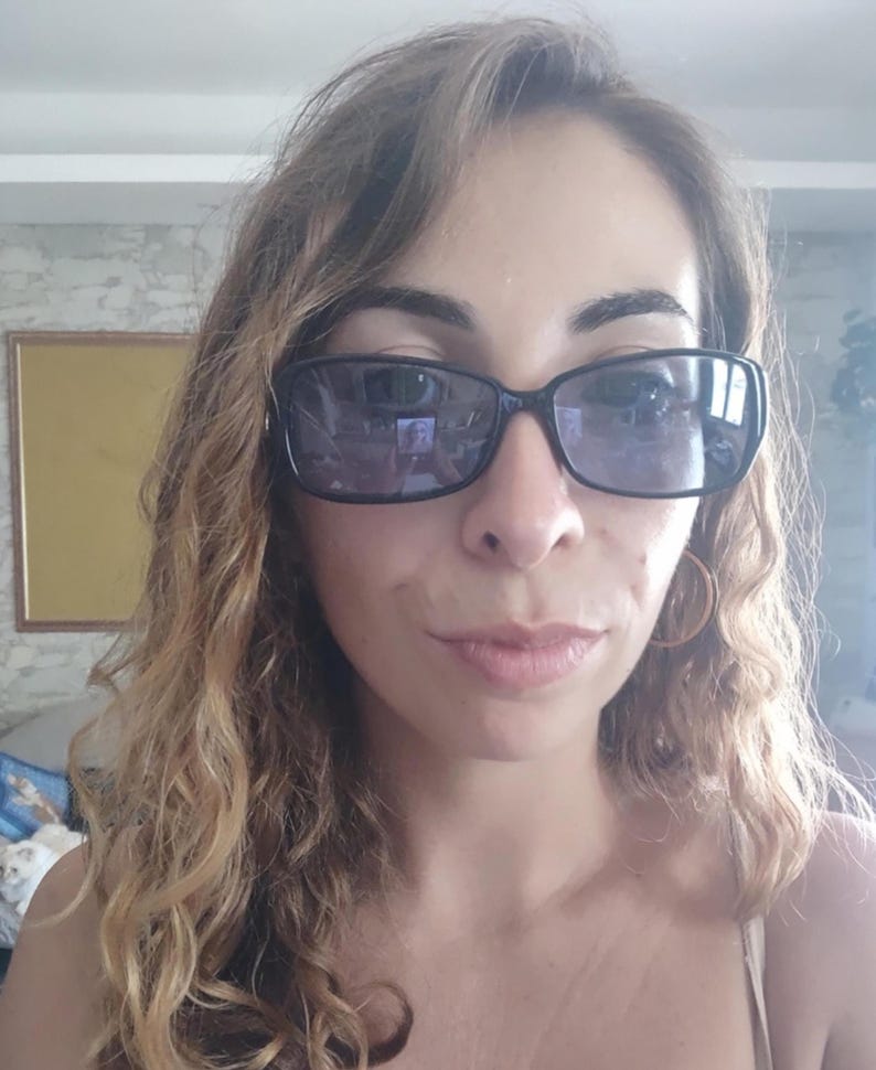 May include: A person wearing black rectangular sunglasses with dark lenses. The person has long, wavy, light brown hair and is wearing gold hoop earrings. The background is a light-colored wall with a framed picture.