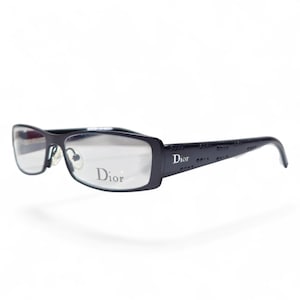 May include: Black rectangular-framed spectacles with clear lenses. The Dior logo is visible on the lens and the temples. The temples have a repeating pattern. The spectacles are set against a white background.
