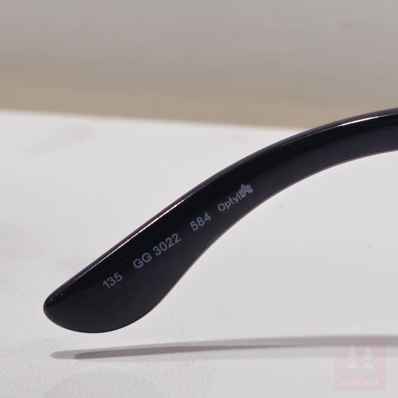 May include: Close-up of a black eyeglass arm. The arm has the following printed on it: "135 GG 3022 584 Optyl". The eyeglass arm is curved and has a glossy finish.