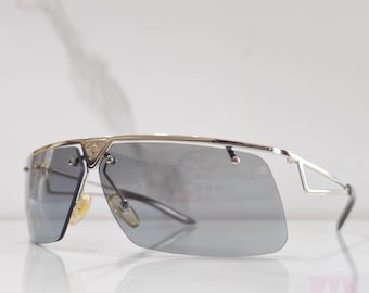 Puede incluir: Gafas de sol aviador plateadas con lentes gris oscuro y montura plateada. Las gafas tienen un diseño único con un pequeño triángulo en el lateral de la montura.