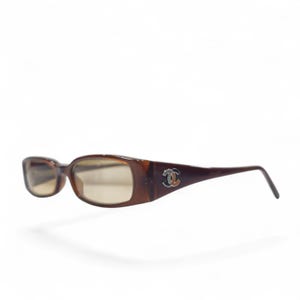 May include: Rectangular sunglasses with a translucent brown tortoiseshell frame and light brown tinted lenses. The temples feature a silver-toned metal logo consisting of two interlocking C letters on each side of the frame.