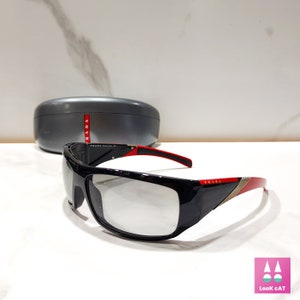 May include: Black sunglasses with red and gold accents. The sunglasses have a rectangular frame and clear lenses. The brand name "PRADA" is visible on the side of the sunglasses.