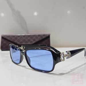 May include: Black rectangular sunglasses with blue lenses. The frames feature a decorative silver-colored embellishment on the temples. A brown case with a diamond pattern is in the background. The sunglasses are on a white surface.