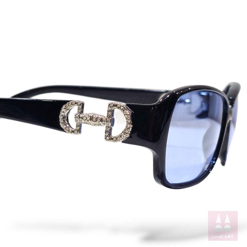 May include: Black sunglasses with blue lenses. The sunglasses feature a decorative silver-colored embellishment on the temples. The lenses are a light blue color. The frame is a glossy black.