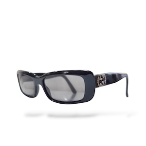 May include: Black rectangular sunglasses with dark tinted lenses and a thick frame. The temples feature a prominent silver-toned circular Gucci logo emblem on the side, connecting the frame front to the arms of the eyewear.