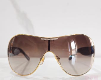 May include: Gold-rimmed sunglasses with gradient brown lenses. The sunglasses have a curved, shield-like design with a small row of rhinestones across the top. The bridge and arms are gold-toned.