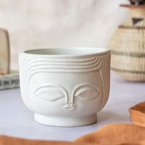 May include: A white ceramic planter with a face design. The planter features a serene face with closed eyes and a subtle lip. The design includes etched lines that create the appearance of hair. The planter is a decorative item for plants.