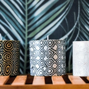 May include: Three decorative candles with geometric patterns. The candles are on a wooden shelf in front of a dark green and black tropical leaf patterned background. The candles are black and white, black and gold, and white with a gray pattern.