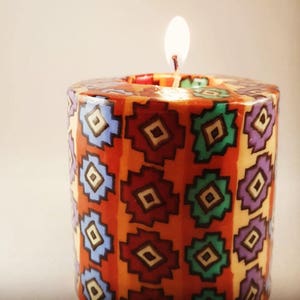 May include: A lit pillar candle with a vibrant geometric pattern. The candle features a burnt orange base, adorned with a repeating design of red, blue, green, and purple diamond shapes. The flame glows brightly, and the candle is centered.