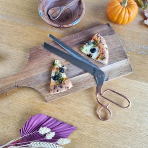 May include: A wooden serving board with two pizza slices, a small pumpkin, and a wooden bowl. A pizza cutter with copper handles and a stainless steel blade is on the board. The pizza has olives and pesto.