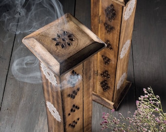 Puede incluir: Dos porta inciensos de madera con intrincados tallados y diseños blancos. El humo se eleva desde la parte superior de los soportes, que están sobre una superficie de madera.