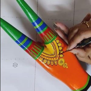 Pode incluir: Close-up de um objeto decorativo de madeira pintada. O objeto &eacute; laranja com detalhes em verde, azul, vermelho e amarelo. O artista est&aacute; adicionando detalhes com um pincel fino. O objeto tem um design sim&eacute;trico com padr&otilde;es florais e geom&eacute;tricos.