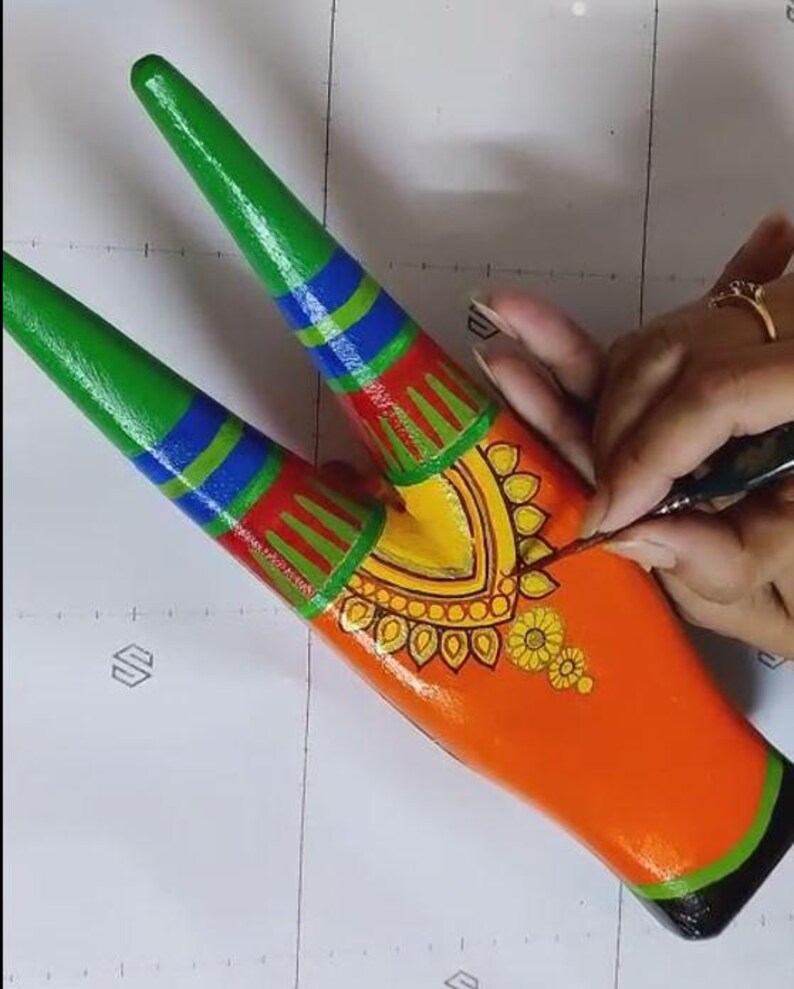 Pode incluir: Close-up de um objeto decorativo de madeira pintada. O objeto &eacute; laranja com detalhes em verde, azul, vermelho e amarelo. O artista est&aacute; adicionando detalhes com um pincel fino. O objeto tem um design sim&eacute;trico com padr&otilde;es florais e geom&eacute;tricos.