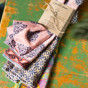 Pu&ograve; includere: Un mucchio di quadrati di tessuto colorati con un'etichetta di carta marrone che recita "SARI GIFT WRAP RECYCLED naamsari.com Ridurre le discariche, conservare l'energia, ridurre l'inquinamento dell'aria e dell'acqua, risparmiare le risorse naturali".