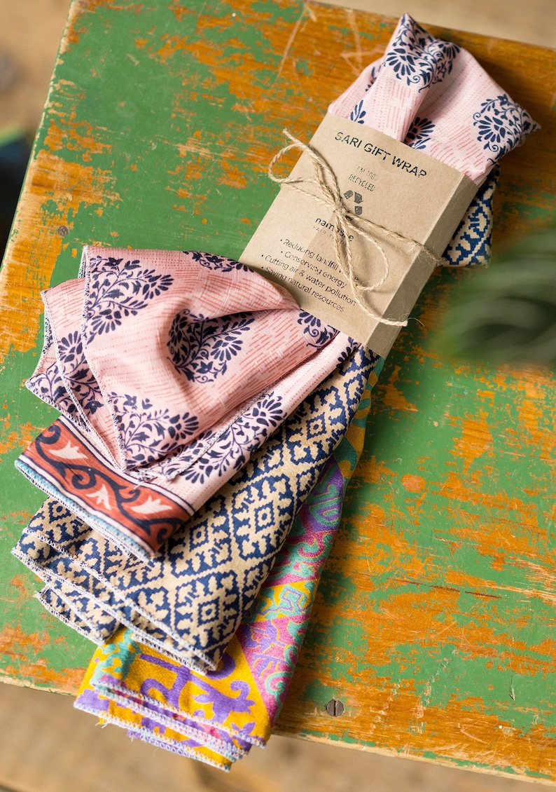 Pu&ograve; includere: Un mucchio di quadrati di tessuto colorati con un'etichetta di carta marrone che recita "SARI GIFT WRAP RECYCLED naamsari.com Ridurre le discariche, conservare l'energia, ridurre l'inquinamento dell'aria e dell'acqua, risparmiare le risorse naturali".