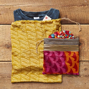 May include: Two fabric drawstring bags, one yellow with a leaf pattern and one with a multi-colored striped top and a pink and orange bottom with a wave pattern. The bags are on a wooden surface.