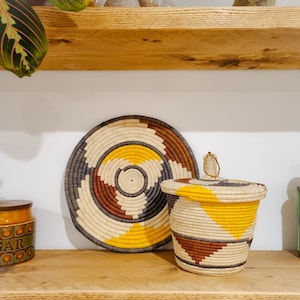 May include: A wooden shelf displays a woven basket with a lid, a round woven plate, and a jar labelled "SUGAR". The basket features yellow, brown, and cream geometric patterns. The plate has a similar design. A green glass jar is also visible.