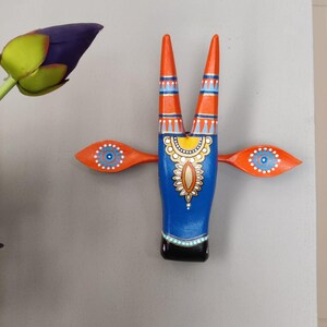 Pode incluir: Uma cabe&ccedil;a de touro de madeira pintada com chifres e asas laranja. O corpo do touro &eacute; azul com intrincados padr&otilde;es amarelos e brancos. As asas apresentam um design de olho azul e branco. Os chifres s&atilde;o laranja com listras azuis e brancas.