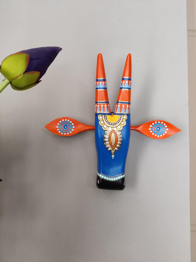 Pode incluir: Uma cabe&ccedil;a de touro de madeira pintada com chifres e asas laranja. O corpo do touro &eacute; azul com intrincados padr&otilde;es amarelos e brancos. As asas apresentam um design de olho azul e branco. Os chifres s&atilde;o laranja com listras azuis e brancas.
