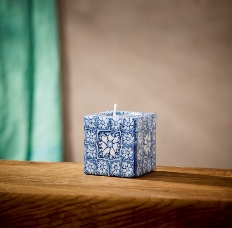 May include: A white candle with a blue and white patterned design. The candle is square shaped and has a single wick.