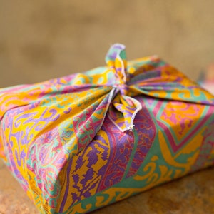 Pu&ograve; includere: Un involucro regalo in tessuto colorato con un motivo paisley in tonalit&agrave; di giallo, rosa, viola e verde. Il tessuto &egrave; legato con un nodo in cima.