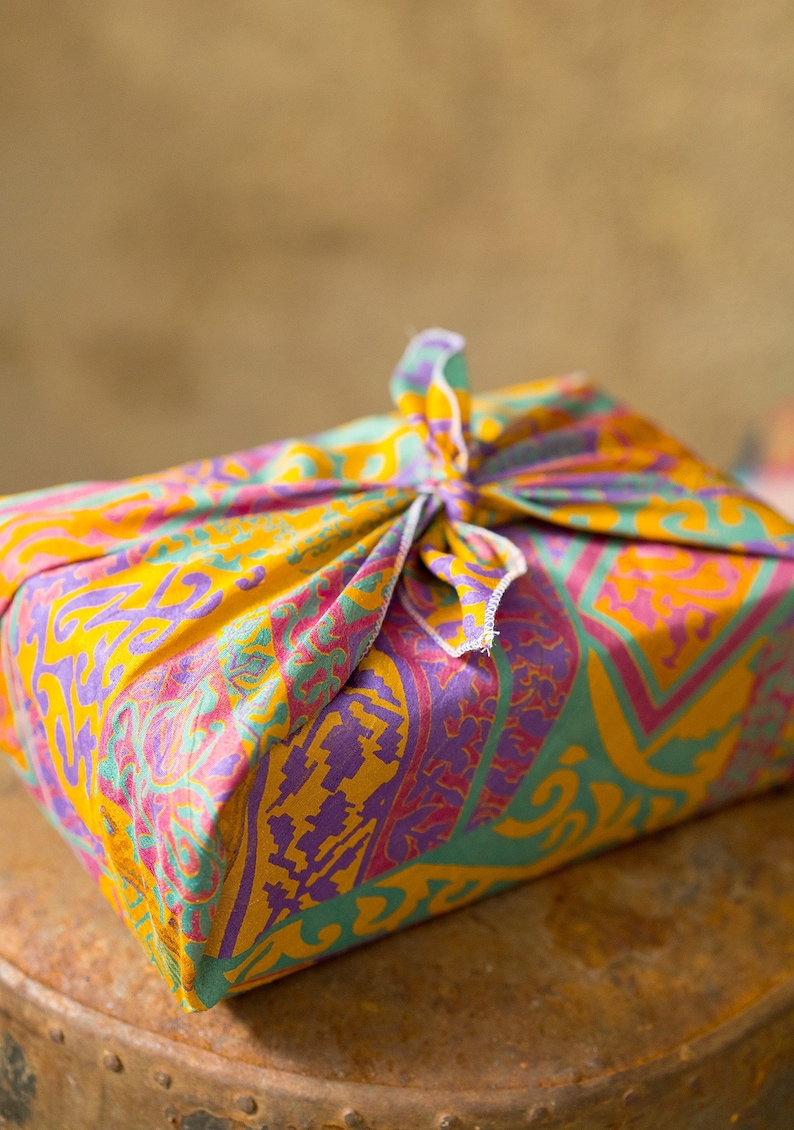 Pu&ograve; includere: Un involucro regalo in tessuto colorato con un motivo paisley in tonalit&agrave; di giallo, rosa, viola e verde. Il tessuto &egrave; legato con un nodo in cima.