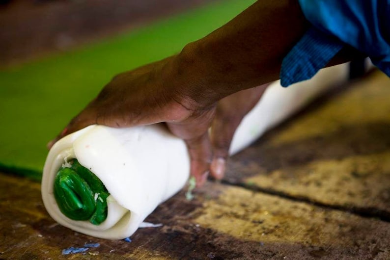 May include: A person rolling a white and green candle with their hand. The candle is being rolled on a wooden surface.