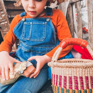 Op de afbeelding: Een jong kind in een blauwe overall en een oranje shirt zit op een houten bankje en houdt een chocolade reep vast die in gouden folie is gewikkeld. Een geweven mand met kleurrijke strepen staat naast het kind.