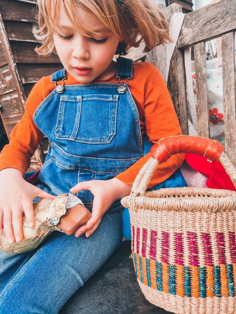 Op de afbeelding: Een jong kind in een blauwe overall en een oranje shirt zit op een houten bankje en houdt een chocolade reep vast die in gouden folie is gewikkeld. Een geweven mand met kleurrijke strepen staat naast het kind.