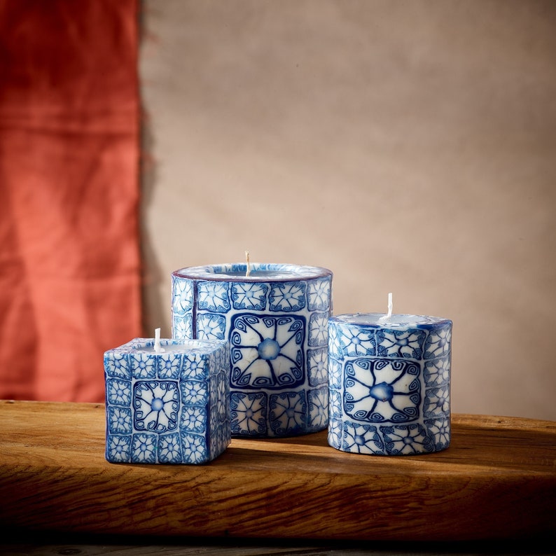 May include: Three decorative candles with blue and white patterned designs. The candles are cylindrical and square in shape and are arranged on a wooden surface.