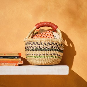 Op de afbeelding: Een geweven mand met een rood leren handvat gevuld met boeken. De mand staat op een wit rek tegen een gele muur.