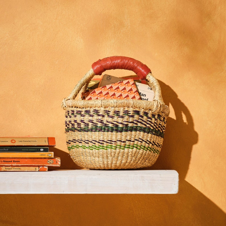 Op de afbeelding: Een geweven mand met een rood leren handvat gevuld met boeken. De mand staat op een wit rek tegen een gele muur.
