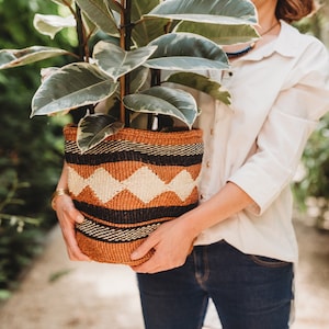 May include: A person holding a large potted plant with variegated leaves in a woven basket with black, brown, and white stripes. The basket is made of natural materials and has a geometric pattern.