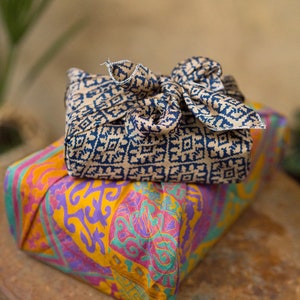 Pu&ograve; includere: Due regali avvolti in tessuto con motivi colorati. Il regalo in alto &egrave; avvolto in un tessuto beige con un motivo geometrico blu scuro. Il regalo in basso &egrave; avvolto in un tessuto giallo con un motivo floreale rosa, viola e verde.