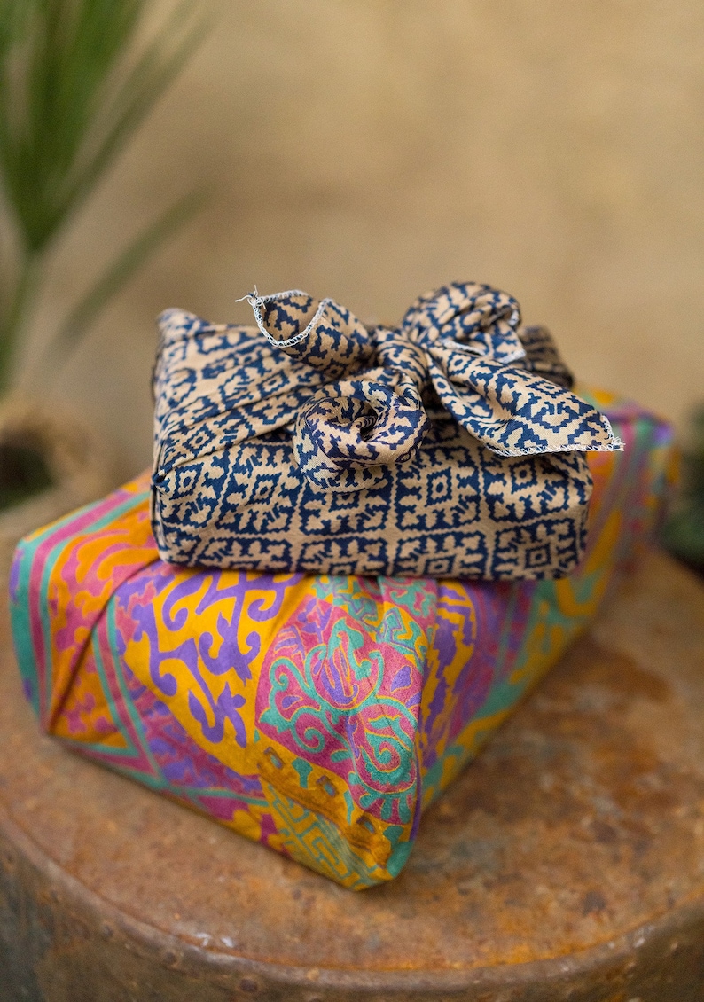 Pu&ograve; includere: Due regali avvolti in tessuto con motivi colorati. Il regalo in alto &egrave; avvolto in un tessuto beige con un motivo geometrico blu scuro. Il regalo in basso &egrave; avvolto in un tessuto giallo con un motivo floreale rosa, viola e verde.